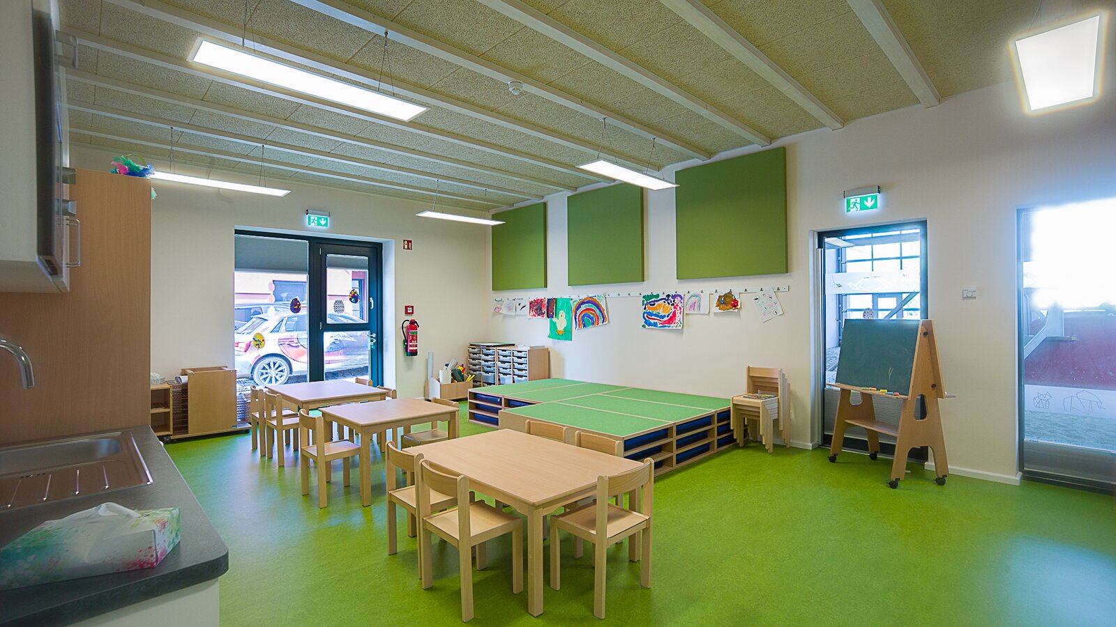 Gruppenraum einer Kindertagesstätte mit grünem Boden und Tischen und Stühlen aus Holz